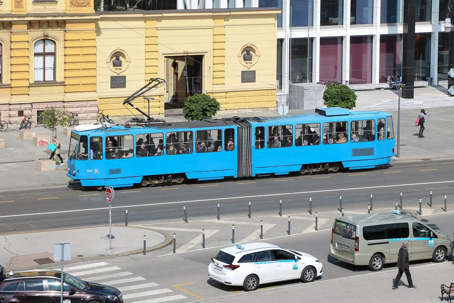 Plavi zagrebački tramvaj pokraj žute zgrade, snimljen odozgo, a na ulici ispod su taksiji.