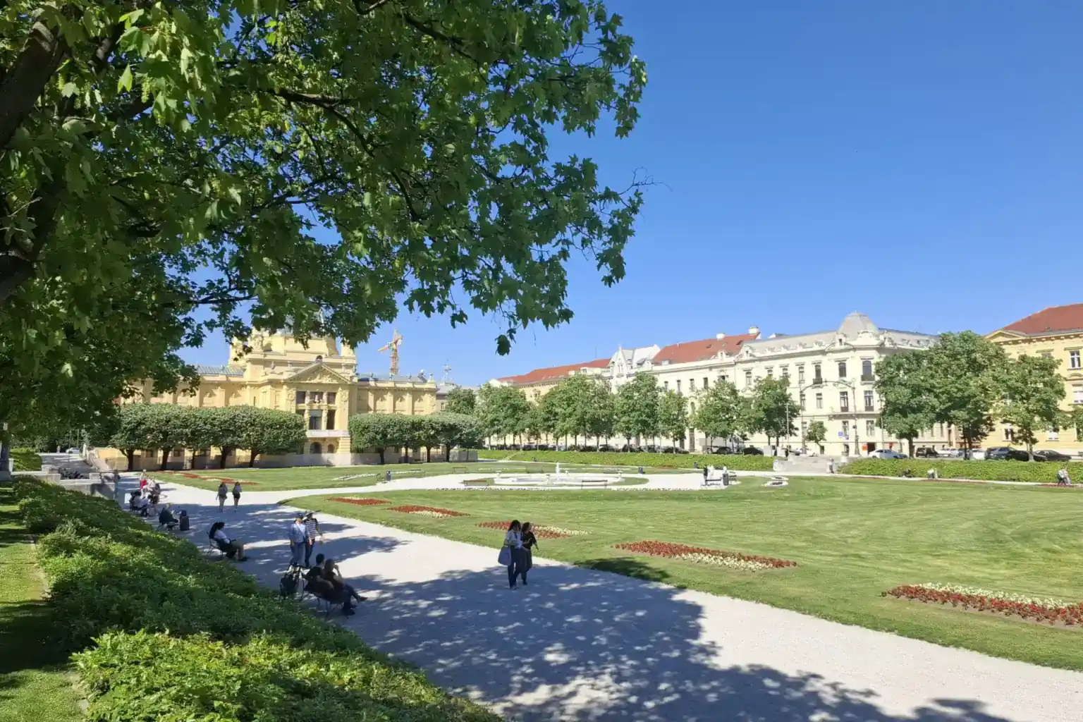 Sunčani park na Trgu kralja Tomislava u Zagrebu s travnjacima, cvjetnim gredicama, šetačima i žutim Umjetničkim paviljonom iza drveća.