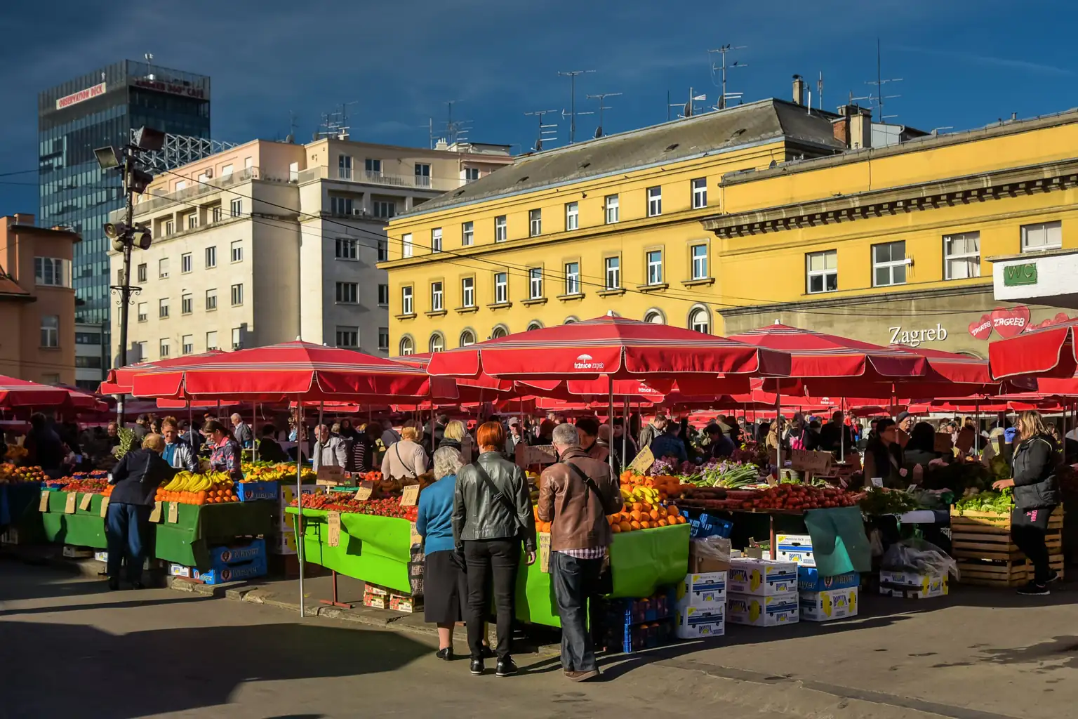 Tržnica Dolac u Zagrebu s crvenim suncobranima nad štandovima s voćem i povrćem i kupcima ispred žutih zgrada.