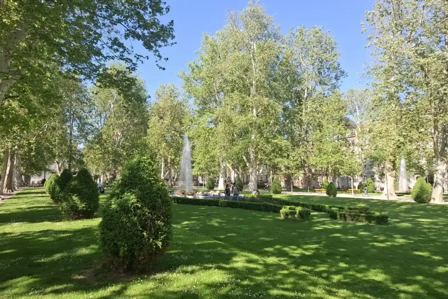 Park Zrinjevac u Zagrebu s fontanom među visokim platanama, šišanim živicama i sunčanim travnjacima.